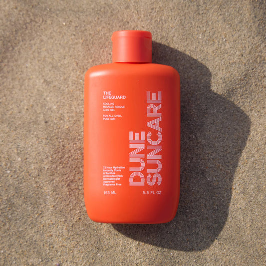 Red bottle of sunscreen that reads 'DUNE SUNCARE THE LIFEGUARD' and care information laying on top of sand