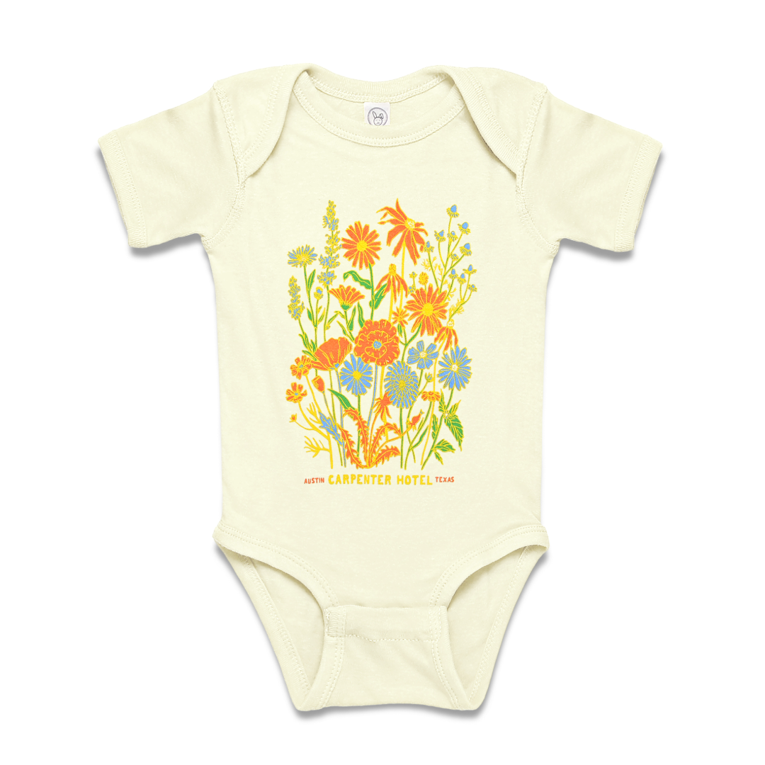 A beige baby onesie with a colorful floral pattern and the text 'Carpenter Hotel austin texas' printed on it.