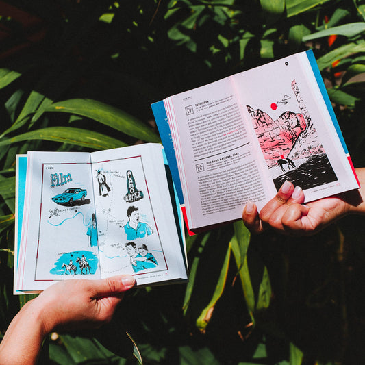 Two people's hands holding open Texas field guides to different pages. One is open to a page about famous filming locations with blue and white illustrations and the other one is open to a page about state and national parks with a red, black and white illustration. There are plant fronds in the background of photo