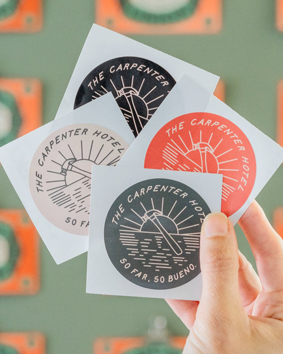 A hand holding four round stickers with a sun motif and the text 'THE CARPENTER HOTEL SO FAR SO BUENO'. The stickers come in different colors including red, olive, black, and tan.