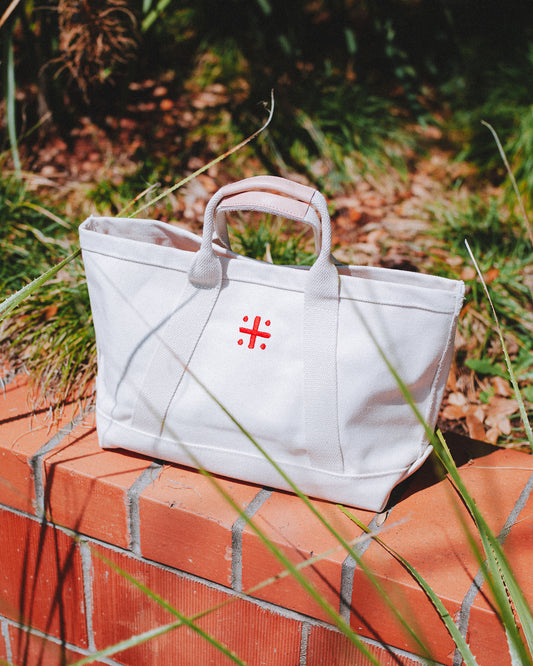 A beige canvas tote bag with leather handles and a red cross logo on the front sitting on brick wall with plants in foreground and background