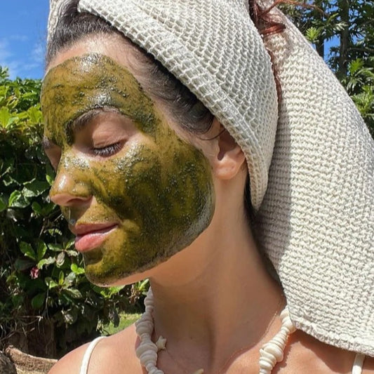 Green face mask rub on person's face while they are wearing a towel on their hair and shell necklace with plants and the sky in the background