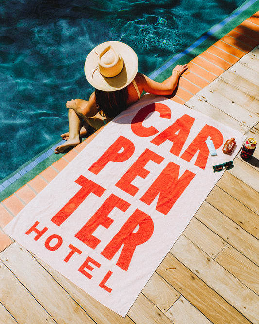 Person sitting by a pool with a light pink and orange pool towel that reads 'CARPENTER HOTEL' in large graphic letters'