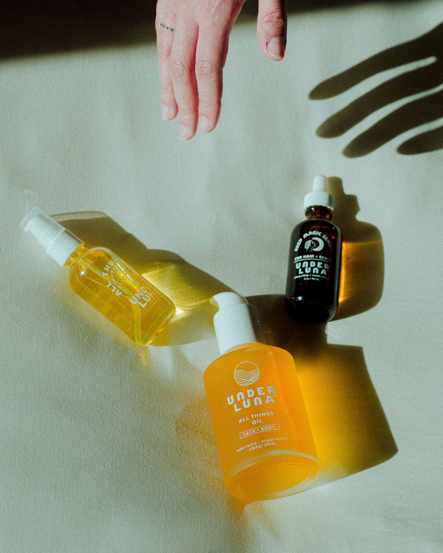 Three bottles of Under Luna skincare products on a light surface with a hand reaching towards them.
