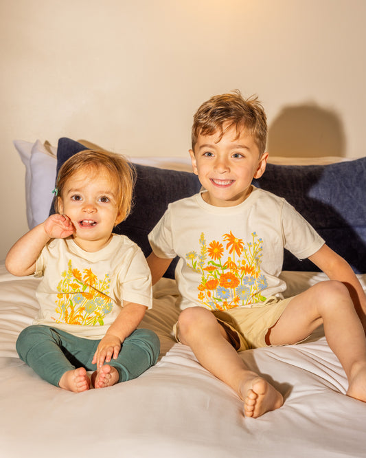A baby and toddler age children sitting on a bed wearing beige kids' shirts with a colorful floral graphic design and text that says ' CARPENTER HOTEL AUSTIN TEXAS' on the chest