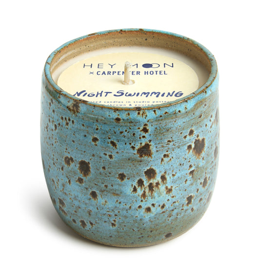 A ceramic candle vessel with a blue and brown speckled design, paper top labeled 'Hey Moon x Carpenter Hotel Night Swimming' with a candle wax insert.