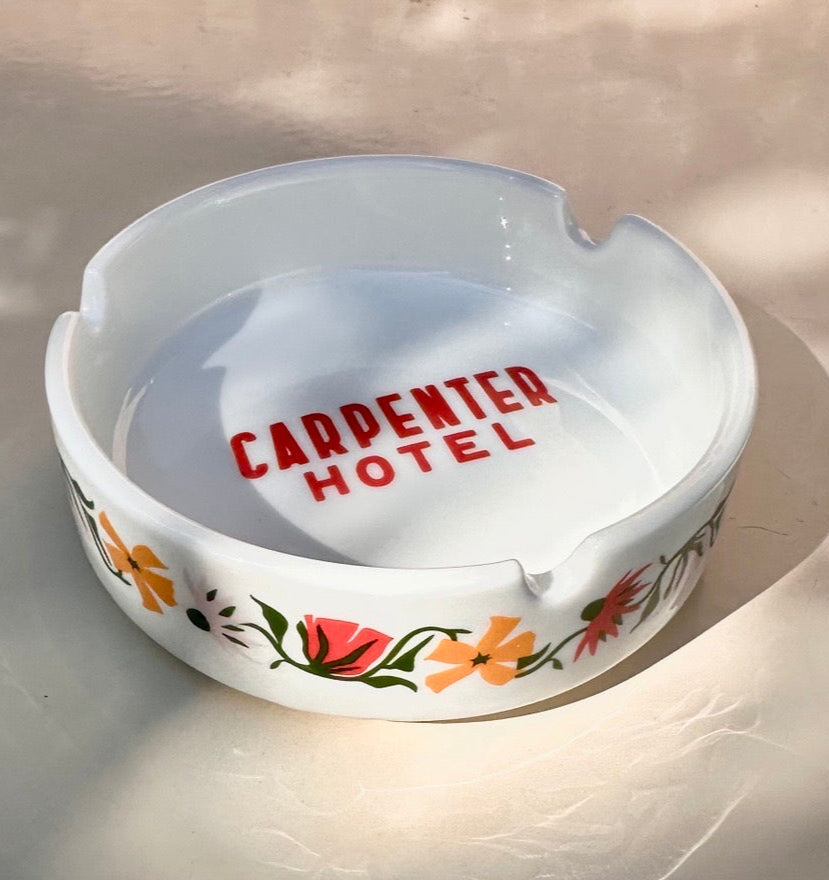 White ceramic ashtray with floral designs on outside and 'CARPENTER HOTEL' text in center bottom sitting on a light background