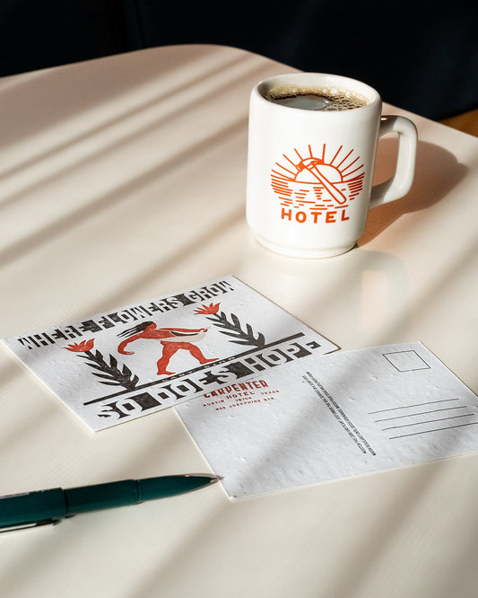 A postcard with letterpress text and illustration, featuring a figure sowing seeds among flowers, with the text 'WHERE FLOWERS GROW SO DOES HOPE - LADY BIRD' pictured with a pen and mug of coffee on top of beige table.