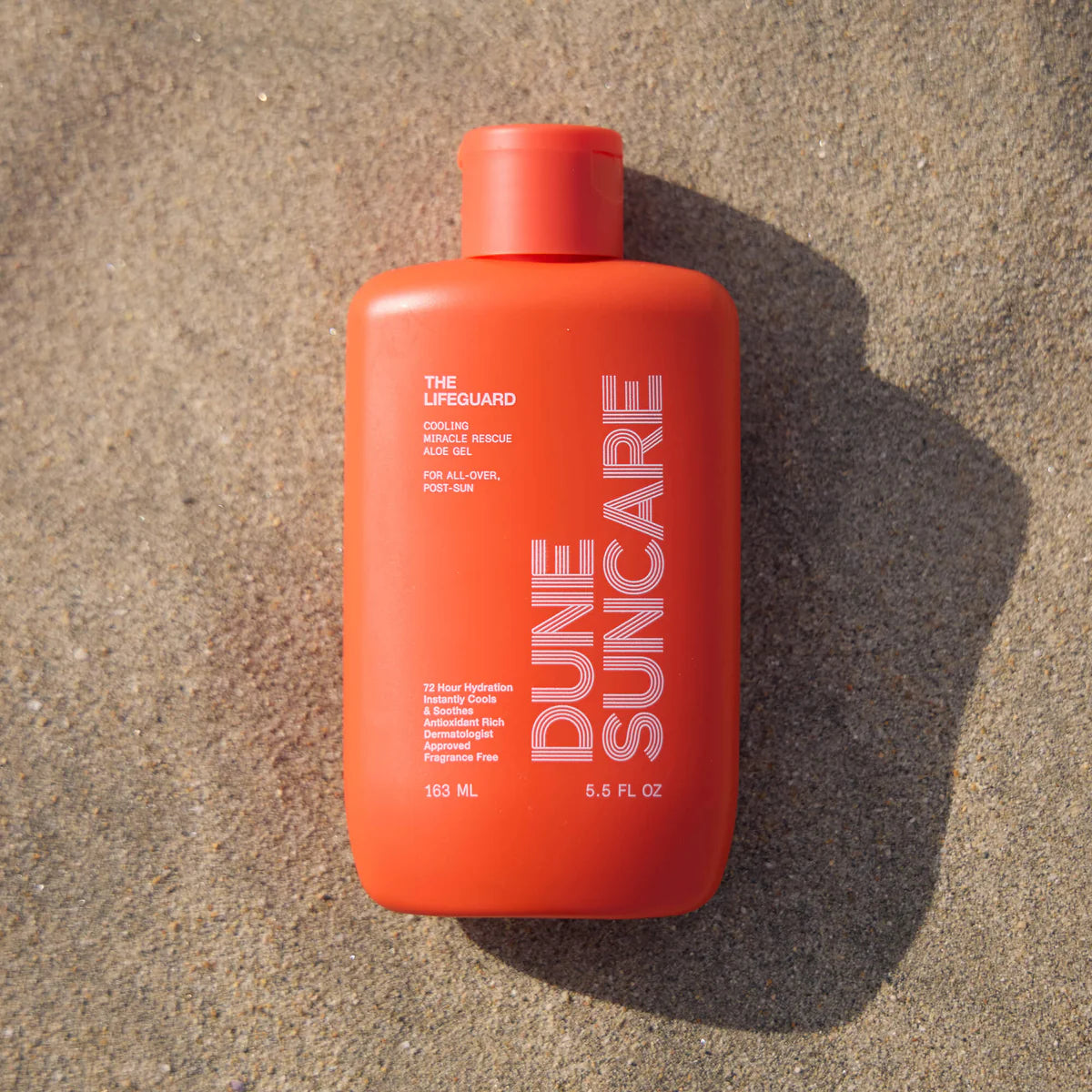 Red bottle of sunscreen that reads 'DUNE SUNCARE THE LIFEGUARD' and care information laying on top of sand