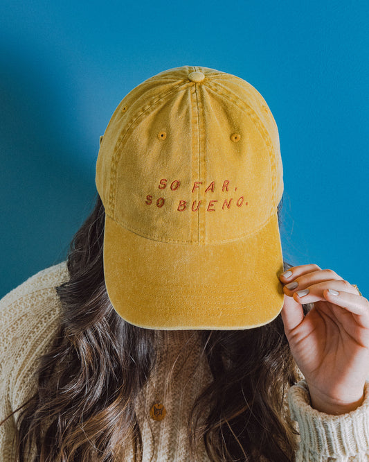Photograph of person wearing yellow 'dad hat' from Carpenter Hotel that says "SO FAR, SO BUENO". Person is wearing a knitted sweater standing in front of a bright blue wall.