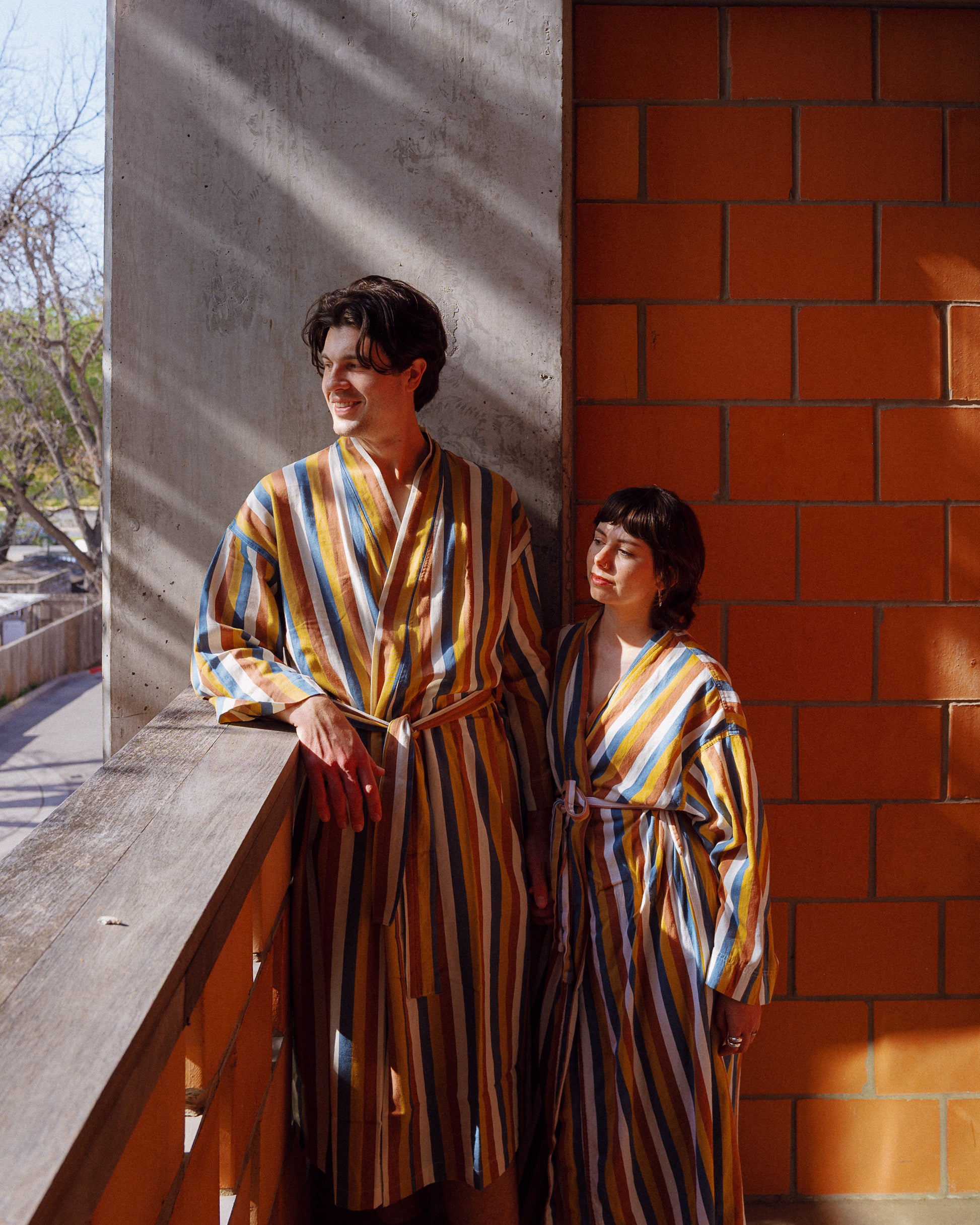 A photograph of two people wearing custom Carpenter Hotel striped robe standing on a hotel balcony made of terracotta, wood, and cement with the sun shining.