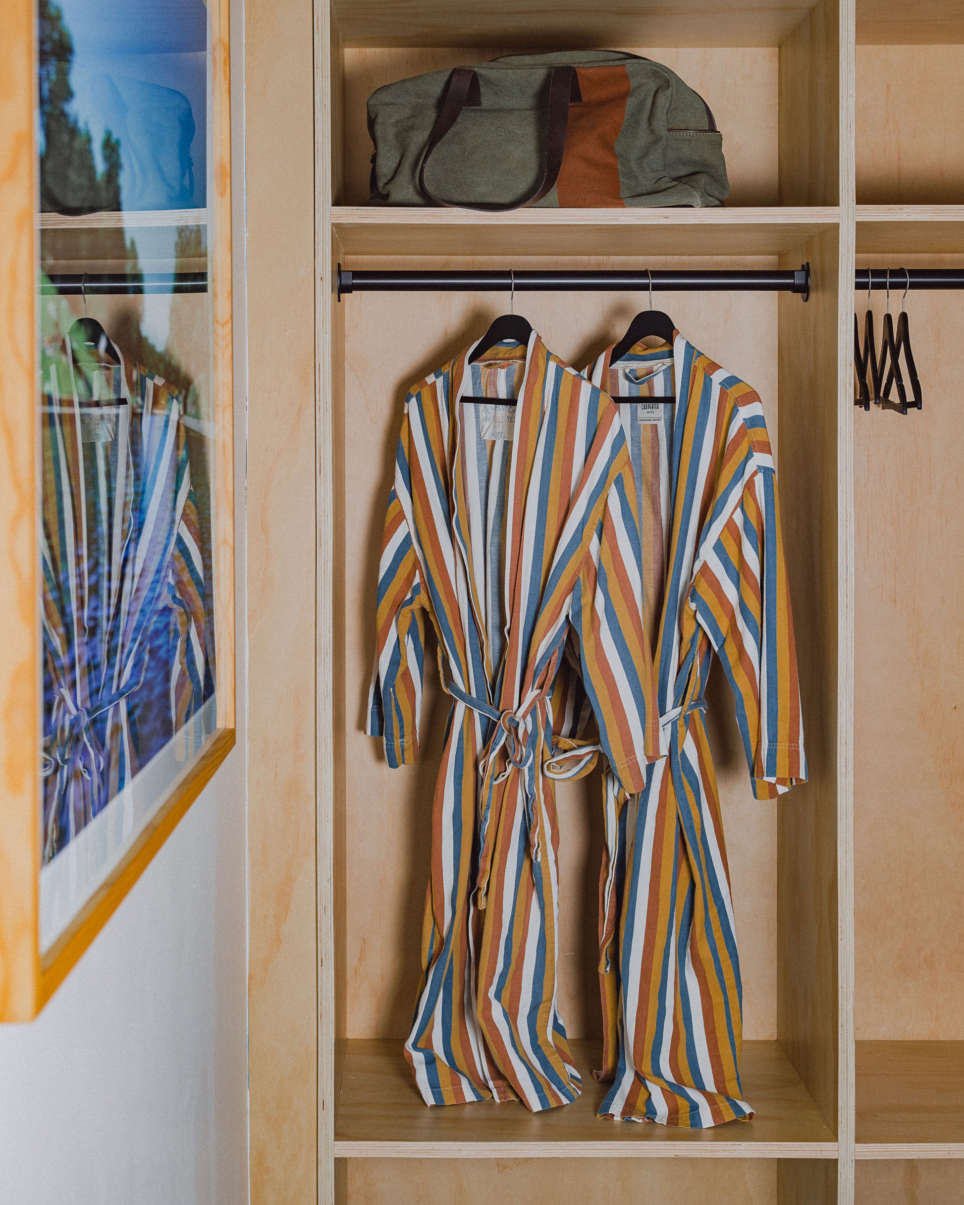 Photograph of two carpenter hotel custom striped robes hanging in cubby closet inside hotel room. Above is a shelf with a duffel bag and a photograph of on opposite wall.