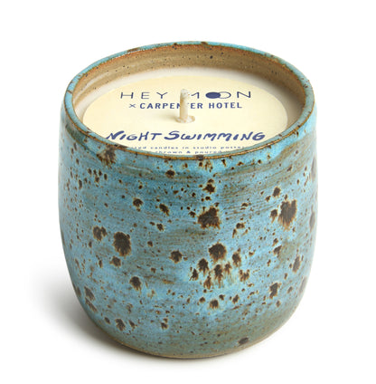 A ceramic candle vessel with a blue and brown speckled design, paper top labeled 'Hey Moon x Carpenter Hotel Night Swimming' with a candle wax insert.