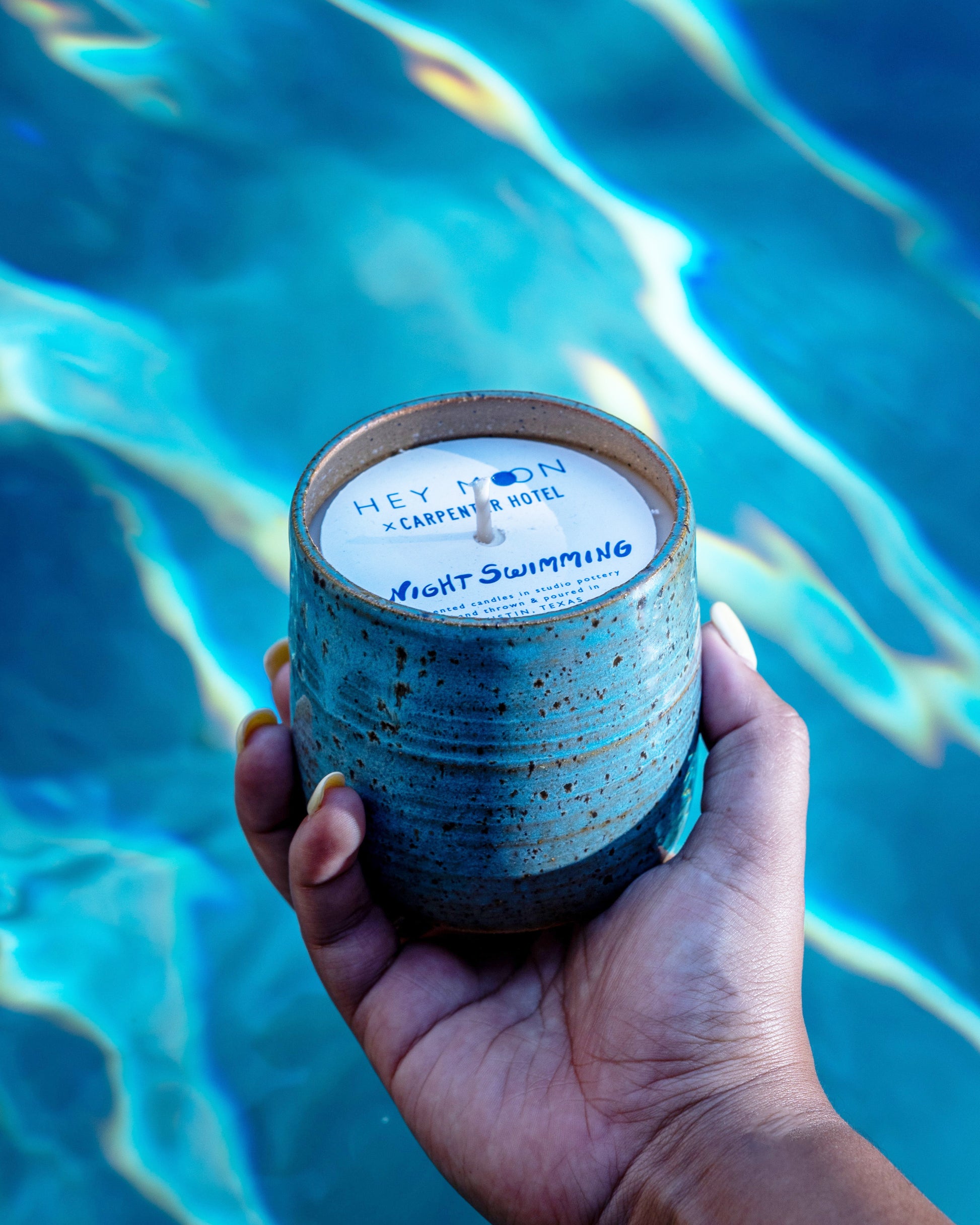 Pool background with hand holding blue with brown speckled ceramic candle vessel. Top paper label reads Hey Moon x Carpenter Hotel Nigh Swimming.