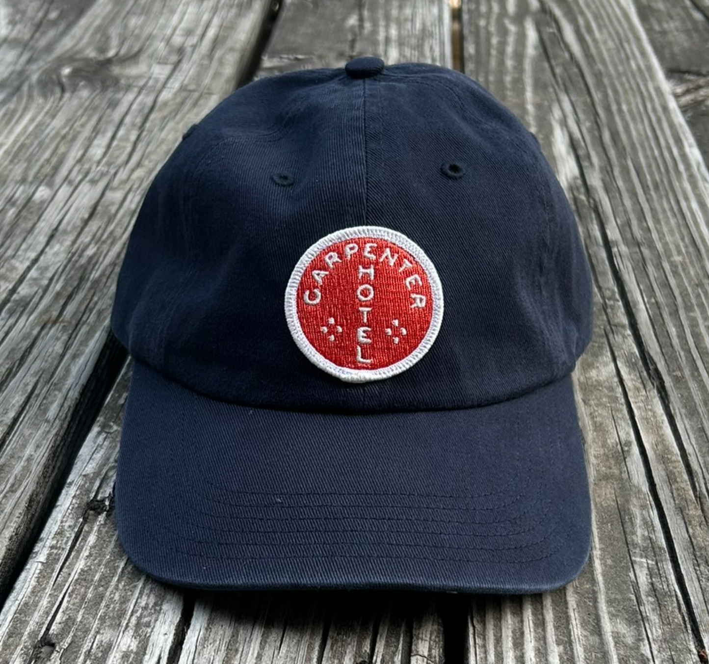 Navy blue hat with orange and white carpenter hotel circle logo patch sitting on gray wooden bench