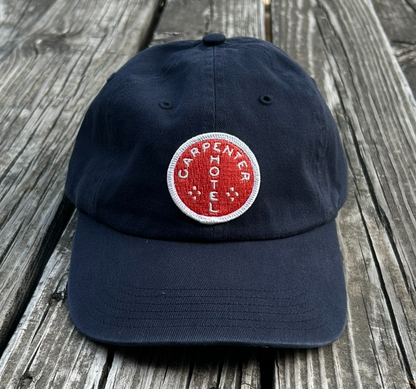 Navy blue hat with orange and white carpenter hotel circle logo patch sitting on gray wooden bench