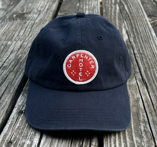 Navy blue hat with orange and white carpenter hotel circle logo patch sitting on gray wooden bench