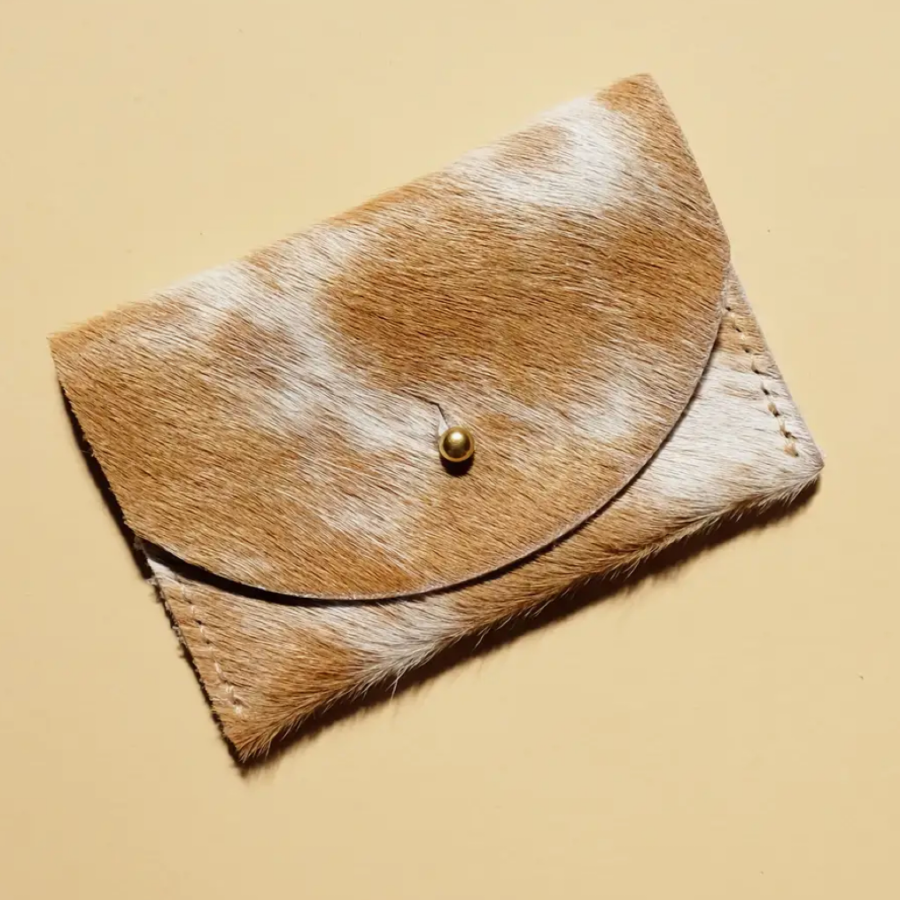 Brown and white cowhide cardholder with gold brass clasp against a light yellow background
