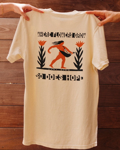 Beige tshirt with black and orange graphic of person carrying seeds with tall flower plant on either side. Text reads 'WHERE FLOWERS GROW SO DOES HOPE' being held by two hands against a wooden background