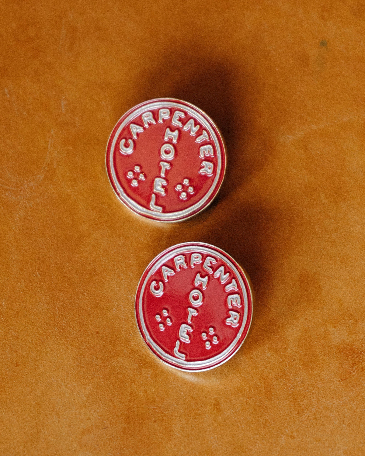 Photograph of two enamel pins with 'CARPENTER HOTEL' circle logo sitting on top of leather
