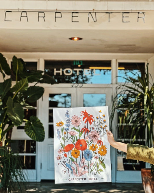 White and glass doorway background with plants on either side with hand holding a colorful poster featuring a variety of illustrated wildflowers with the text 'Austin Carpenter Hotel Texas' at the bottom.