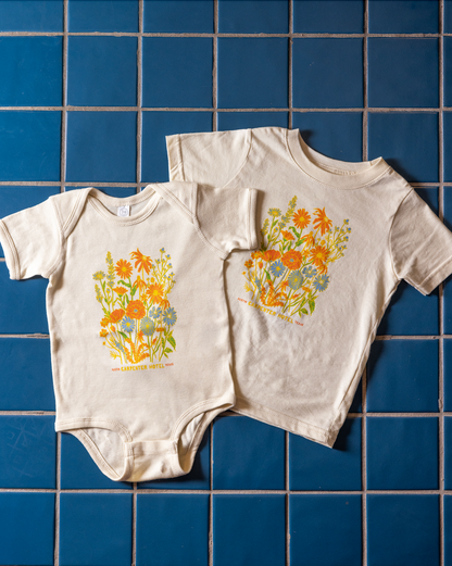 A beige baby onesie and kids t-shirt with a colorful floral pattern and the text 'Carpenter Hotel austin texas' printed on it laying on top of blue tile