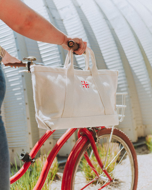A beige canvas tote bag with leather handles and a red cross logo on the front hanging on bicycle handlebars being pushed by a person in front of a metal building