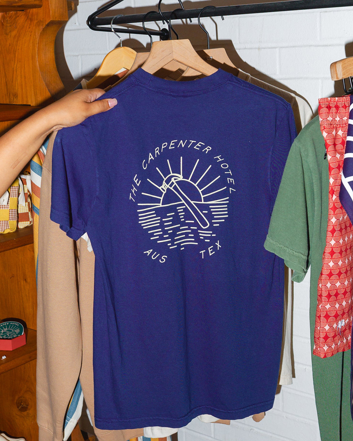 Photograph of navy blue tshirt that says 'THE CARPENTER HOTEL AUS TEX' with line drawing of hammer and sunset in center. The shirt is hanging on a wooden hanger, being held by someone with wooden shelves with other products in the background.