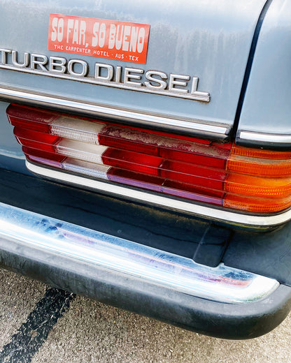 An orange and beige bumper sticker with the text 'SO FAR, SO BUENO THE CARPENTER HOTEL AUST TEX' on the back of light blue vintage car.