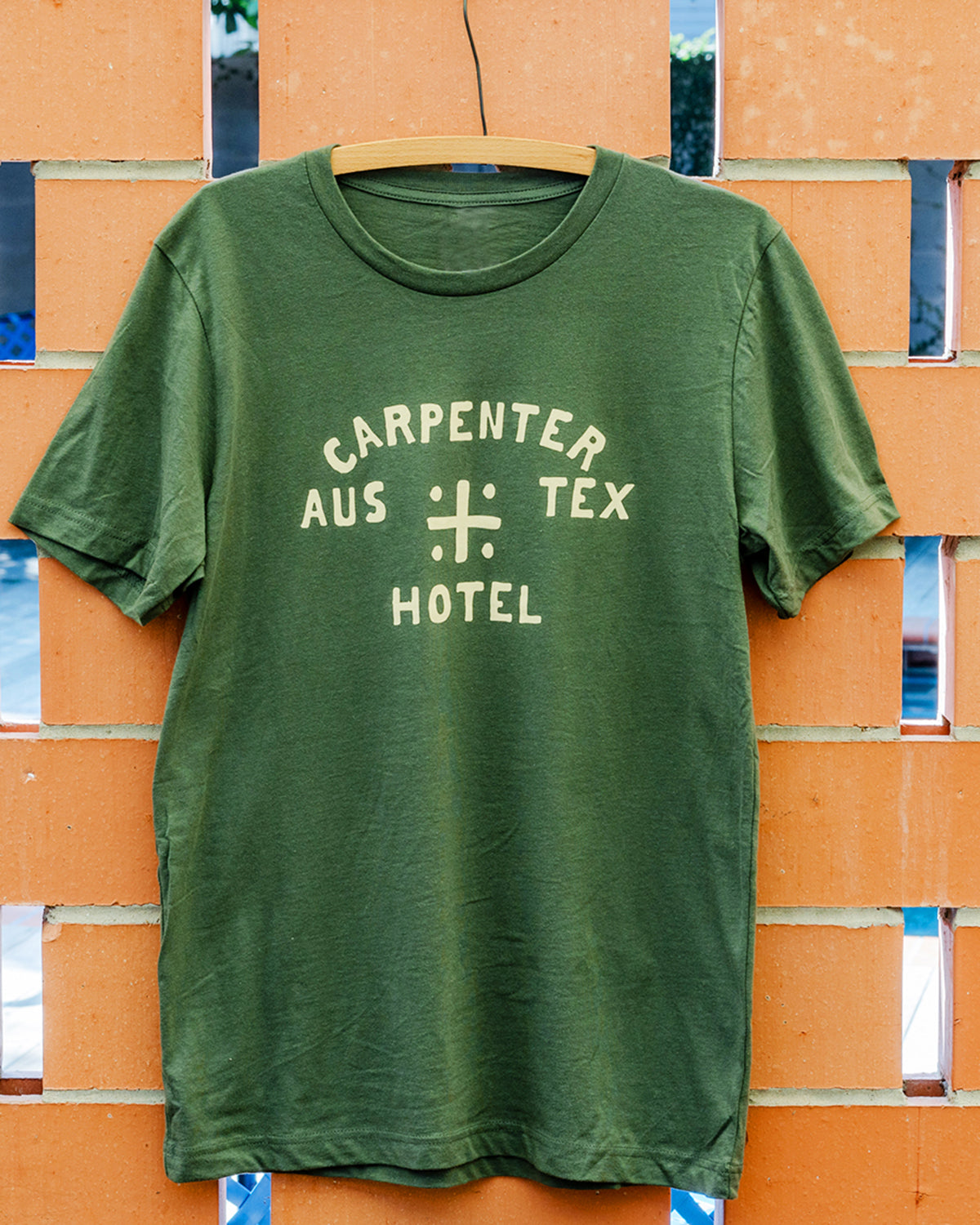 Green tshirt with 'CARPENTER HOTEL AUS TEX' on front in beige graphic. The shirt is hanging on wooden hanger against a terracotta brick background.