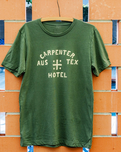 Green tshirt with 'CARPENTER HOTEL AUS TEX' on front in beige graphic. The shirt is hanging on wooden hanger against a terracotta brick background.