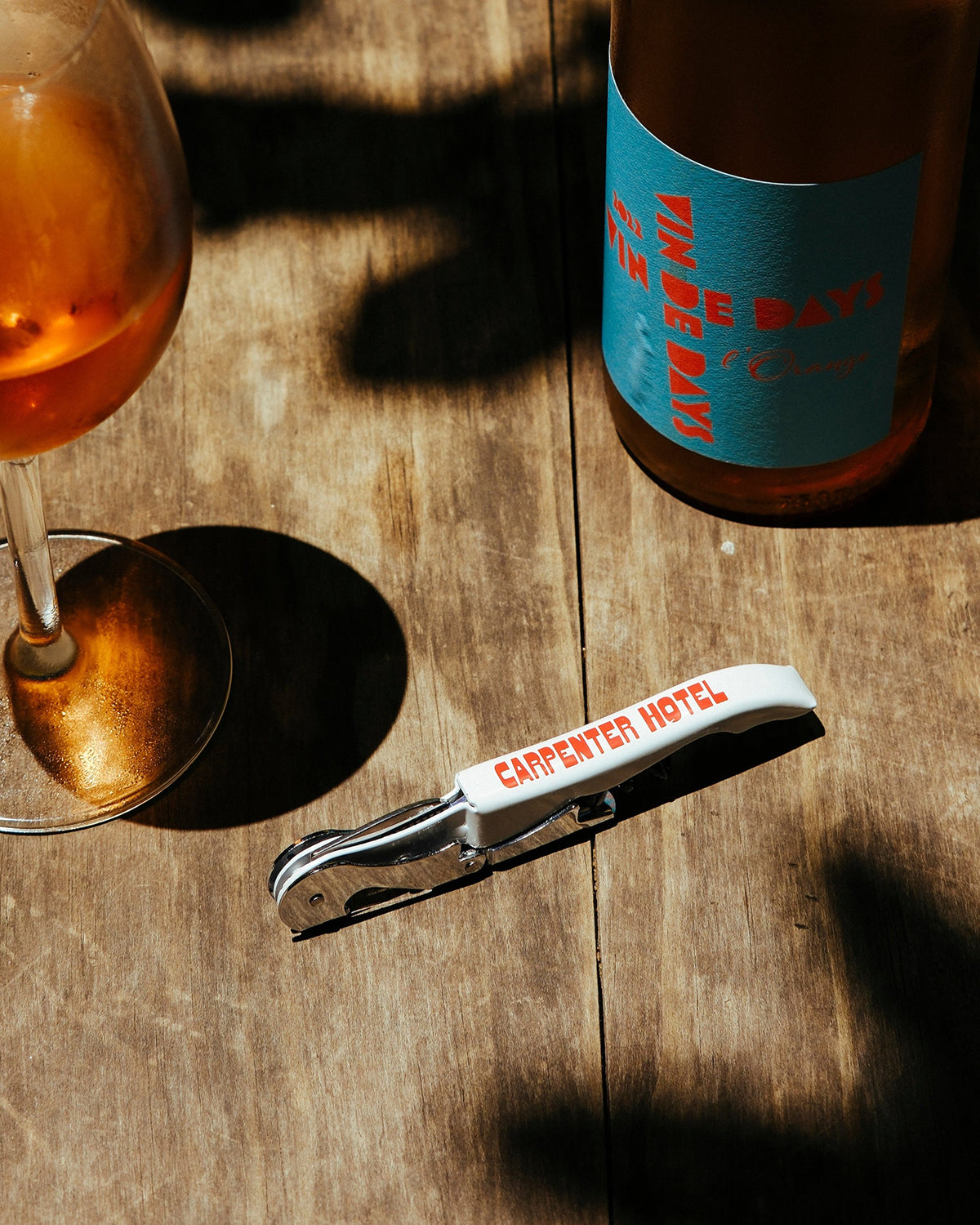 A silver metal wine key with the Carpenter Hotel logo printed in red on its side sitting on a wooden table alongside a glass and bottle of wine