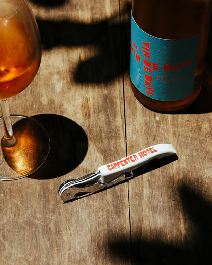 A silver metal wine key with the Carpenter Hotel logo printed in red on its side sitting on a wooden table alongside a glass and bottle of wine