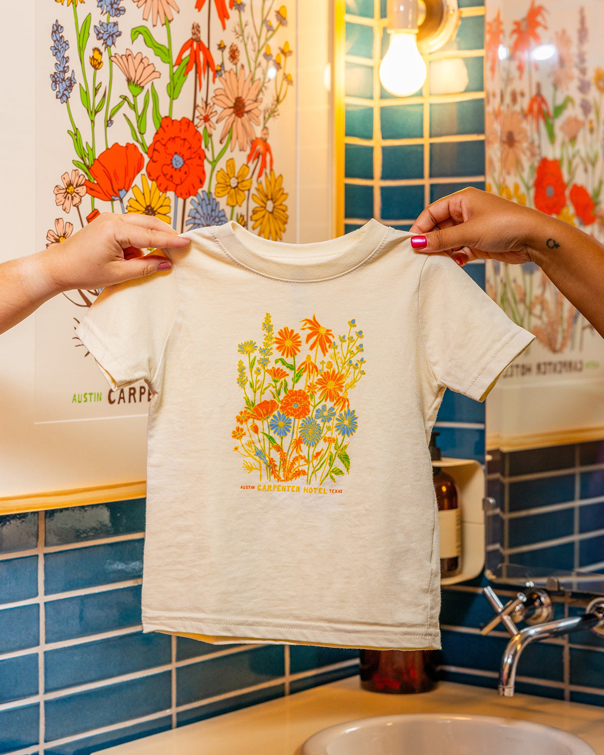 A beige kids' t-shirt with a colorful floral graphic design and text that says ' CARPENTER HOTEL AUSTIN TEXAS' on the chest being held up by two hands with matching poster hanging in background against blue tile.